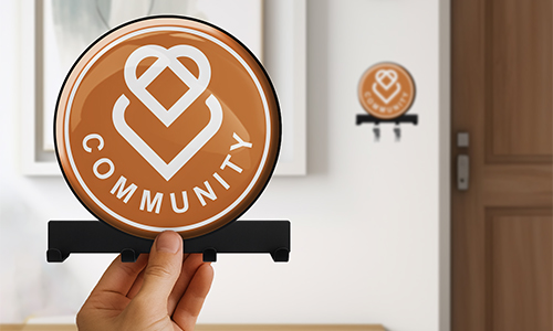 Hand holding circular COMMUNITY sign with heart logo, orange background; same sign mounted on wall near door