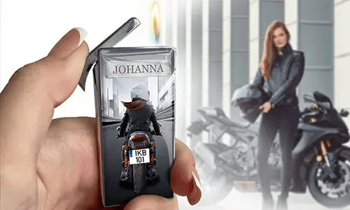 Eine Hand hält ein geöffnetes Feuerzeug mit der Gravur „JOHANNA“ und einem Motorradfahrer-Motiv. Im Hintergrund steht eine Frau in schwarzer Kleidung neben einem Motorrad und hält einen Helm.