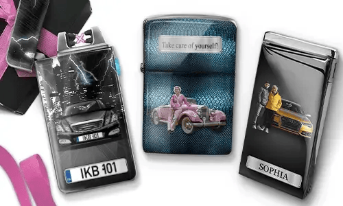 Drei Metallfeuerzeuge mit personalisierten Designs. Das linke Feuerzeug zeigt eine Stadtsilhouette und ein Paar neben einem Auto mit der Aufschrift „BESTE OMA DER WELT“. Das mittlere Feuerzeug stellt ein rosafarbenes Oldtimer-Auto dar, darüber steht „HAB DICH LIEB OMA“. Das rechte Feuerzeug zeigt ein blaues Auto und ein Kennzeichen. Rosafarbene Schleifen verleihen dem Ganzen einen Geschenkcharakter.