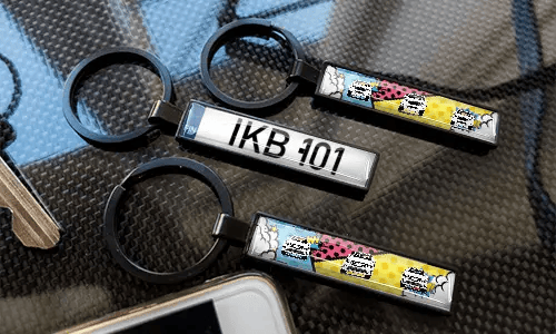 Porte-clés avec plaque d’immatriculation style bande dessinée, exposés sur une surface à texture carbone avec des designs de voitures colorés